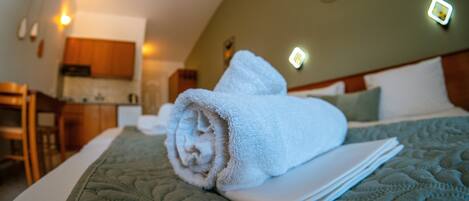 A neatly made bed with a rolled towel and a folded pillow, a wooden headboard, and a kitchen area in the background.