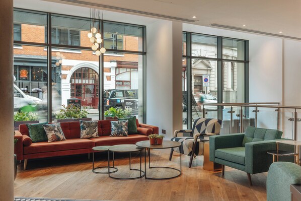 A modern lounge area with a red sofa, green armchairs, and round tables. Large windows offer views of the street and buildings outside.