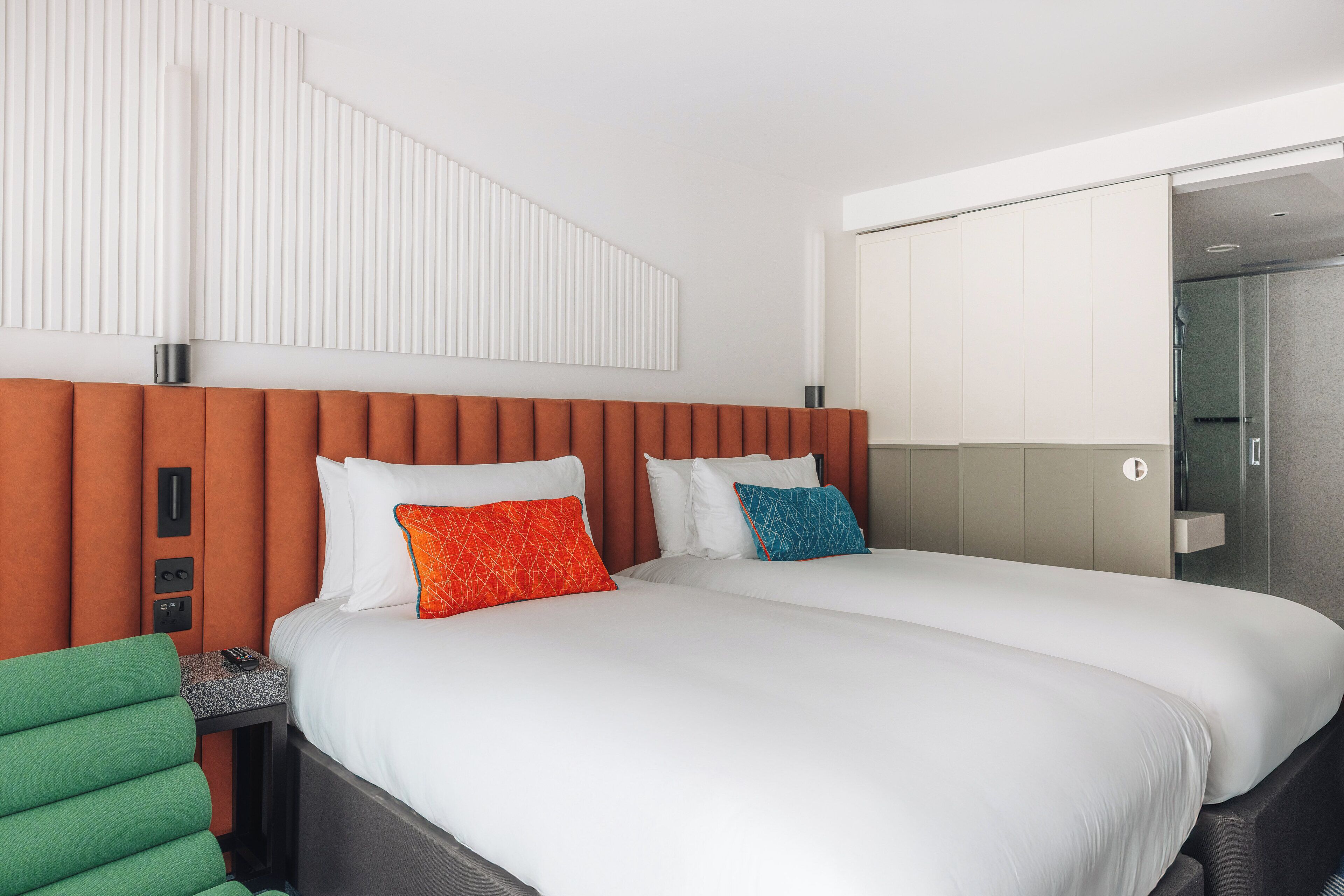 A hotel room with two beds, a green chair, a bedside table with a remote control, and a large mirror.