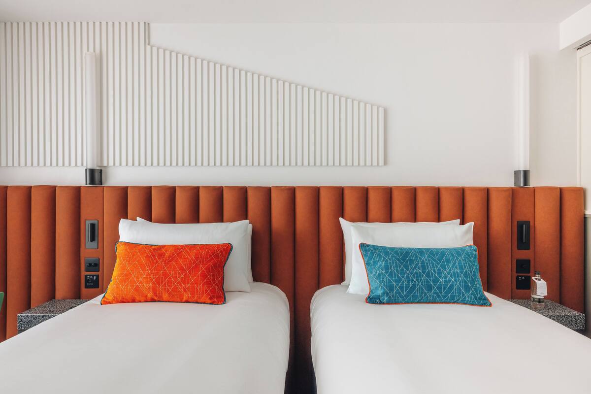 Quarto de hotel com duas camas, cada uma com colchão branco e travesseiro. A cabeceira é laranja com painéis verticais.