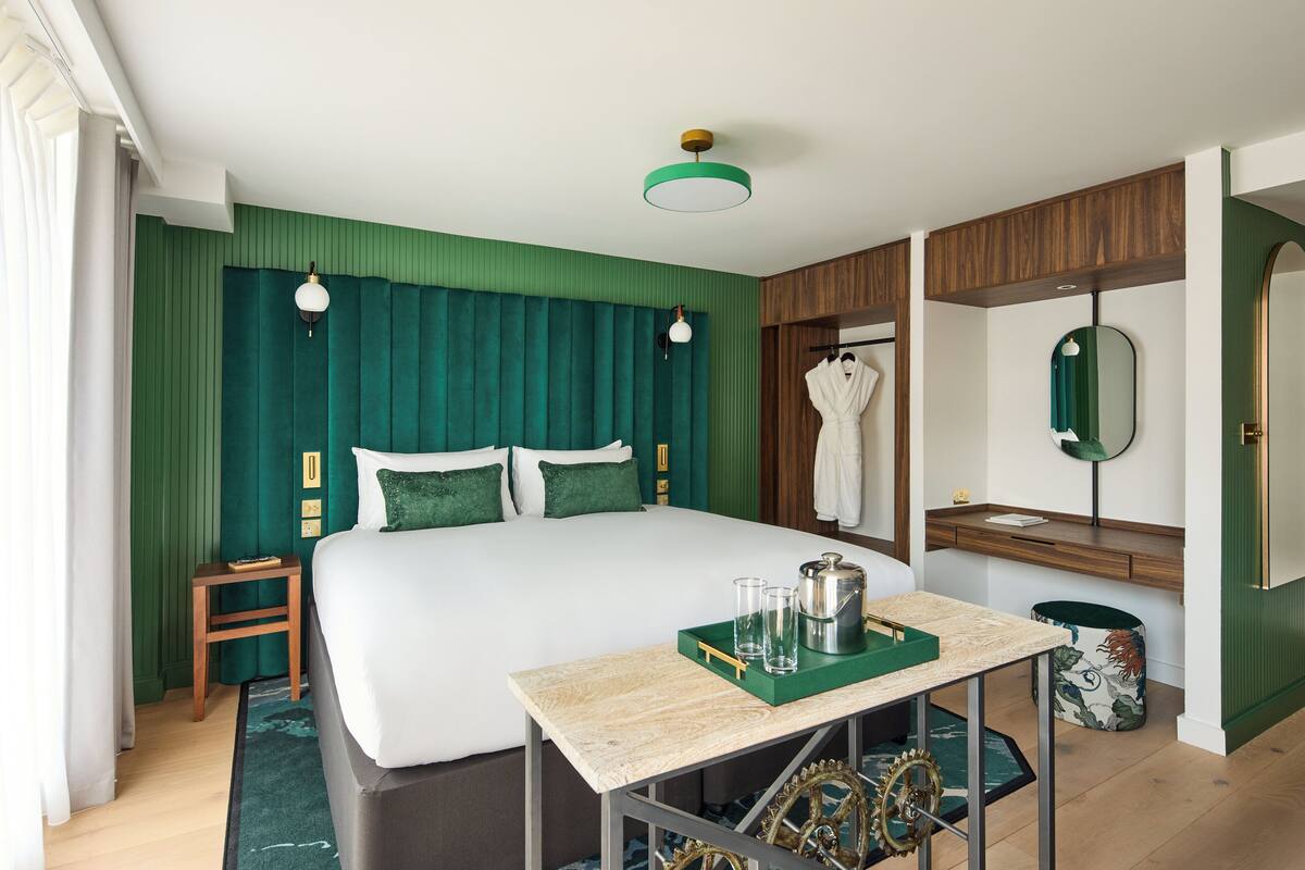 Quarto de hotel com uma cama grande, uma mesinha de cabeceira de madeira, uma mesa de bandeja verde e uma cortina verde.