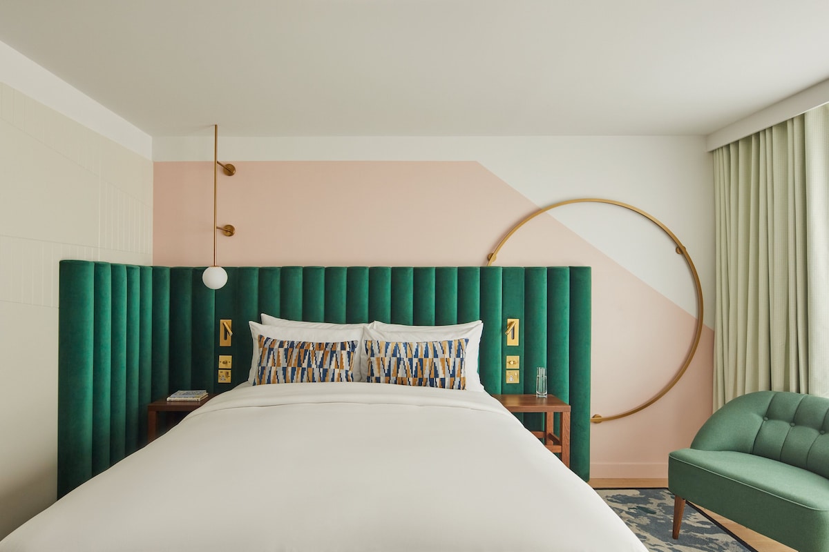 A hotel room with a large bed, a green upholstered headboard, a round mirror, a green armchair, and a bedside table with a lamp.