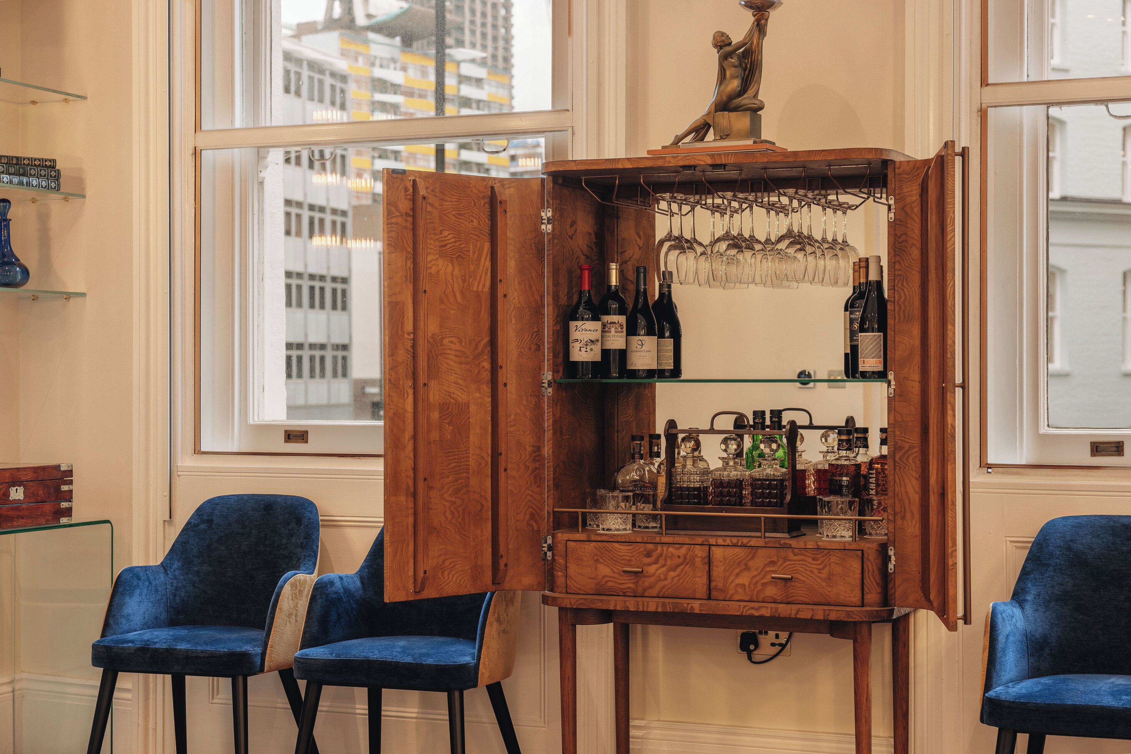 A wooden bar with wine bottles and glasses, two blue chairs, and a statue above the bar.