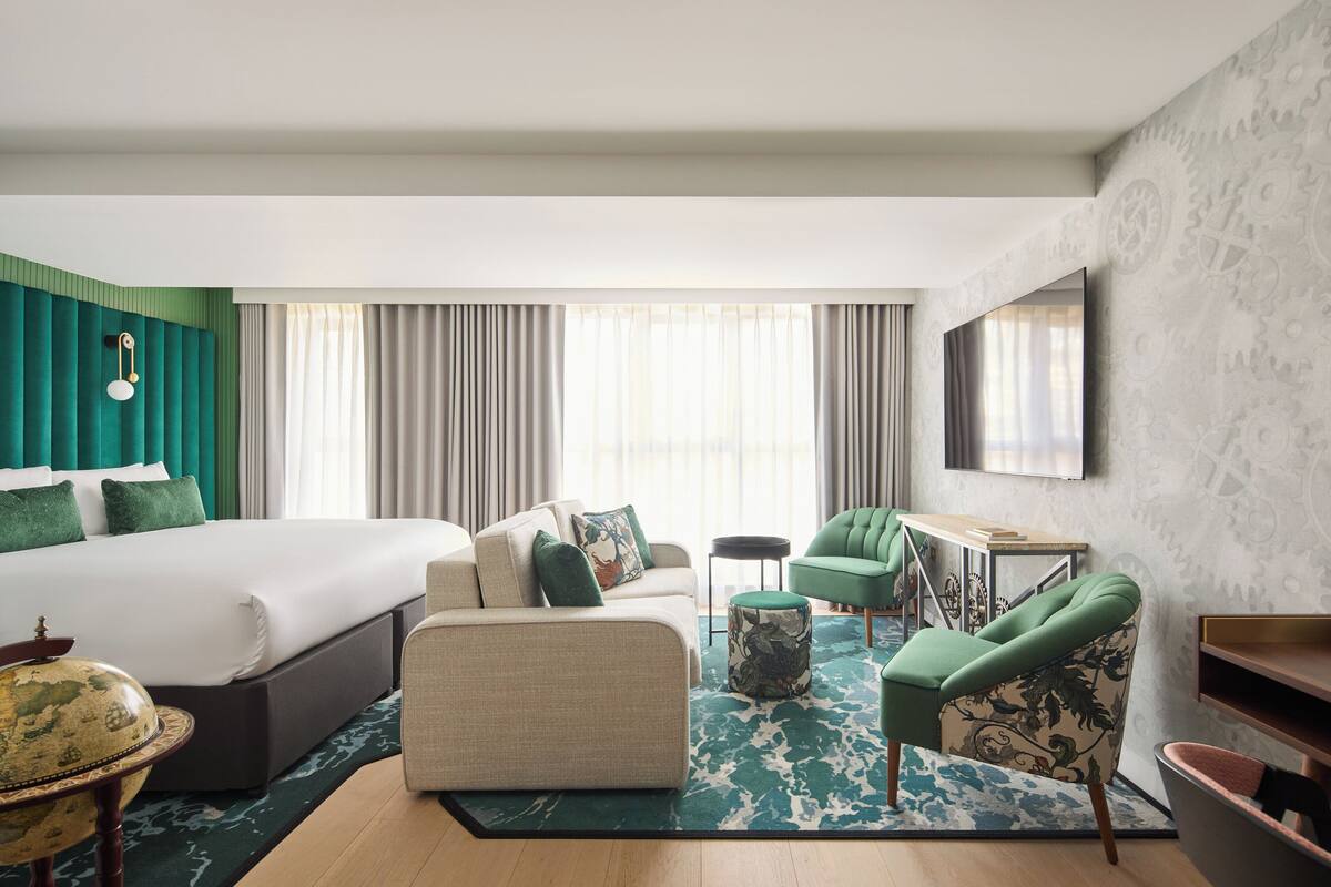 Quarto de hotel com uma cama grande, um sofá, uma cadeira, uma mesa pequena e uma TV fixada na parede.