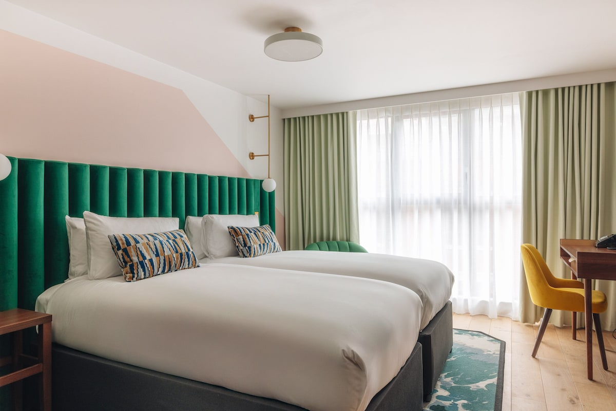 A hotel room with a large bed, a desk, and a chair. There is a window with curtains.