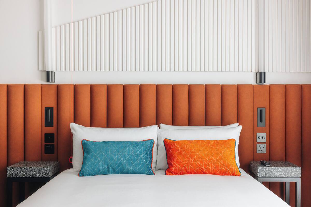 Quarto de hotel com cama, duas almofadas, mesa de cabeceira e painel de controle fixado na parede.