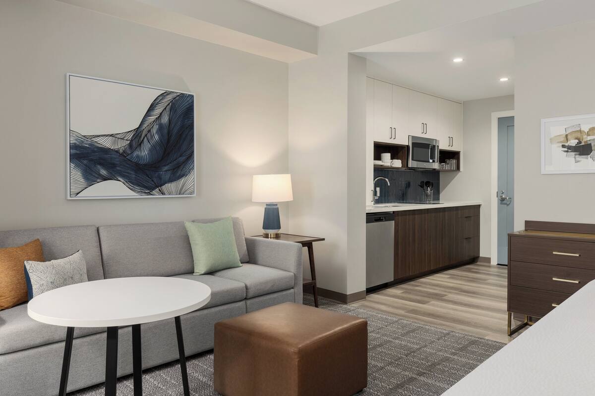Una habitación de hotel moderna con un sofá gris, una mesa redonda blanca, un otoman marrón, una cocina con armarios y electrodomésticos, y un cuadro abstracto enmarcado en la pared.