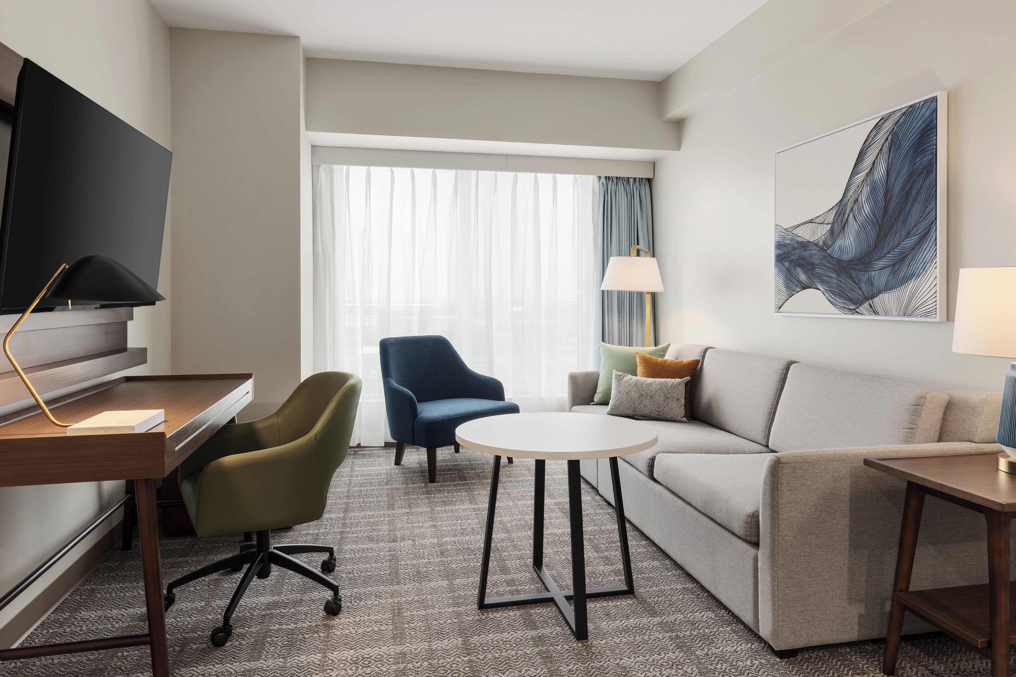 A hotel room with a flat-screen TV, a desk, a green office chair, a blue armchair, a round coffee table, a sofa, and a large abstract painting on the wall.