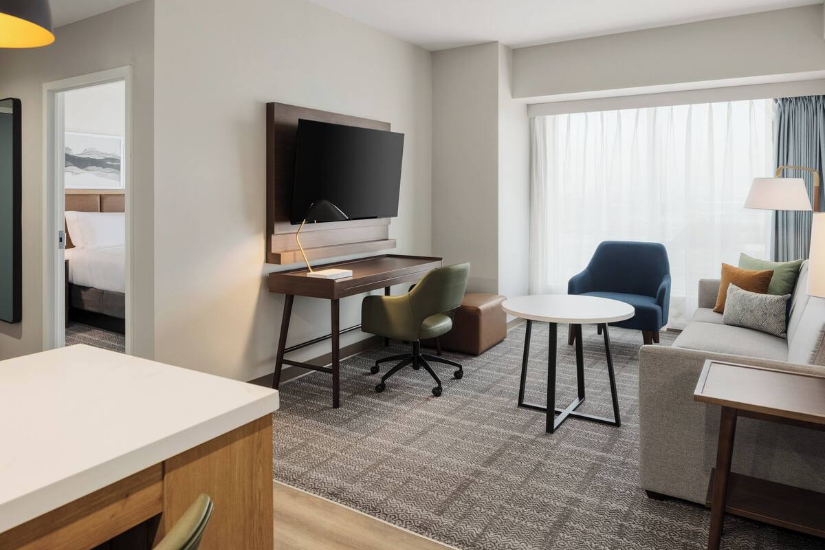 Habitación de hotel con cama, escritorio con televisor de pantalla plana, silla verde, sillón azul, mesita redonda pequeña y sofá con cojines.