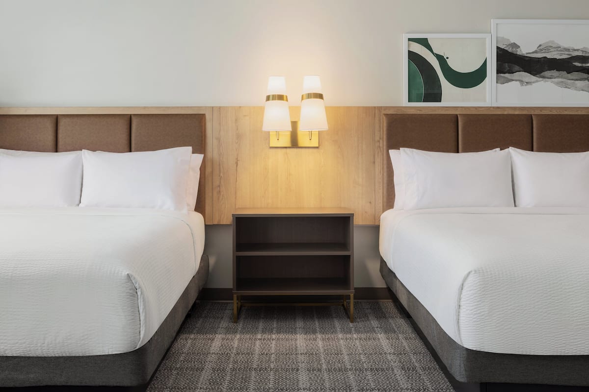 Habitación de hotel con dos camas, una mesita de noche, lámparas fijadas a la pared y una obra de arte en la pared.