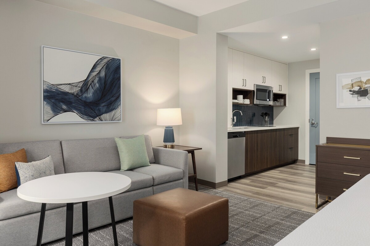 Una habitación de hotel moderna con un sofá gris, una mesa redonda blanca, un otoman marrón, una cocina con armarios y electrodomésticos, y un cuadro abstracto enmarcado en la pared.