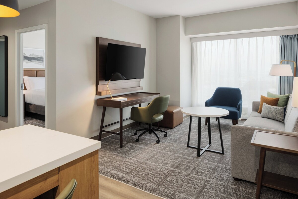 Habitación de hotel con cama, escritorio con televisor de pantalla plana, silla verde, sillón azul, mesita redonda pequeña y sofá con cojines.