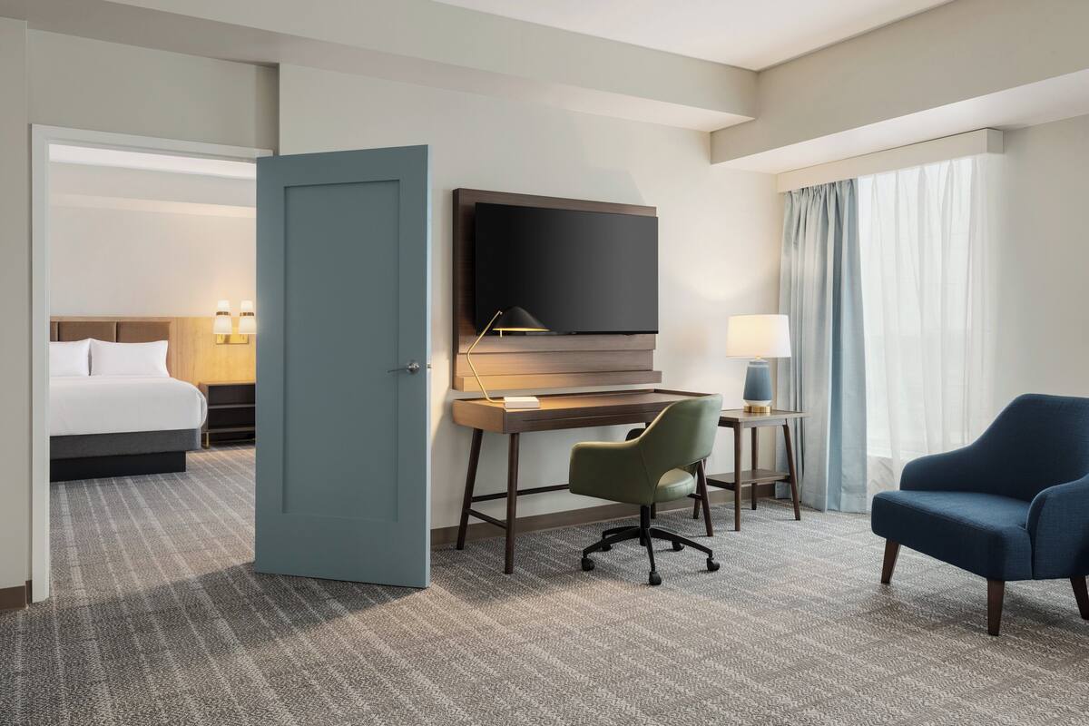 Habitación de hotel con cama, escritorio con televisor, silla, lámpara y ventana con cortinas.