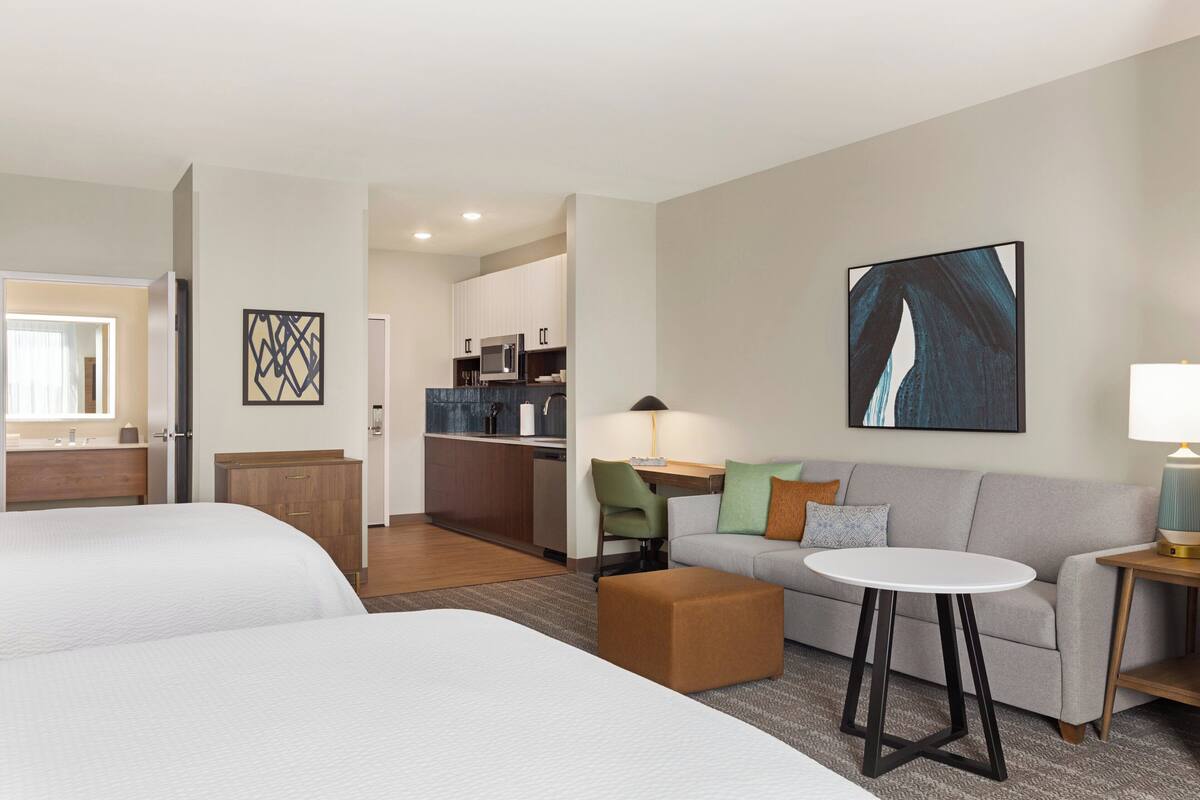 A hotel room with two beds, a sofa, a small table, and a kitchenette in the background.