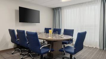 A conference room with a round table, six blue chairs, a flat-screen TV, and a window with sheer curtains.