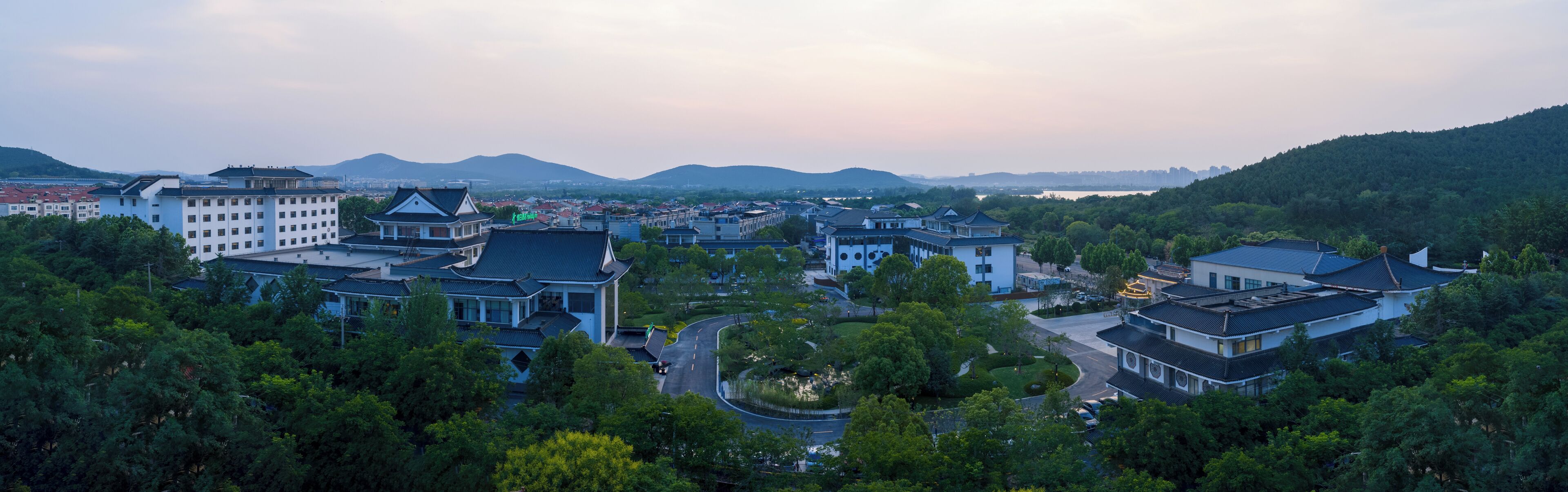 A traditional Chinese architectural style building complex with multiple structures, surrounded by lush greenery and a hilly landscape in the background.