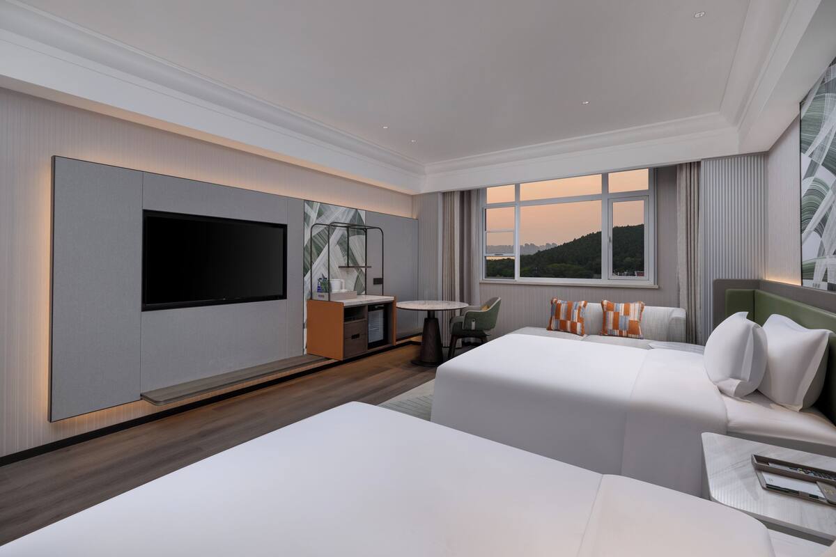 Ein modernes Hotelzimmer mit Flachbildfernseher, Schreibtisch mit Stuhl, zwei Betten und einem Fenster mit Blick auf die Stadt.