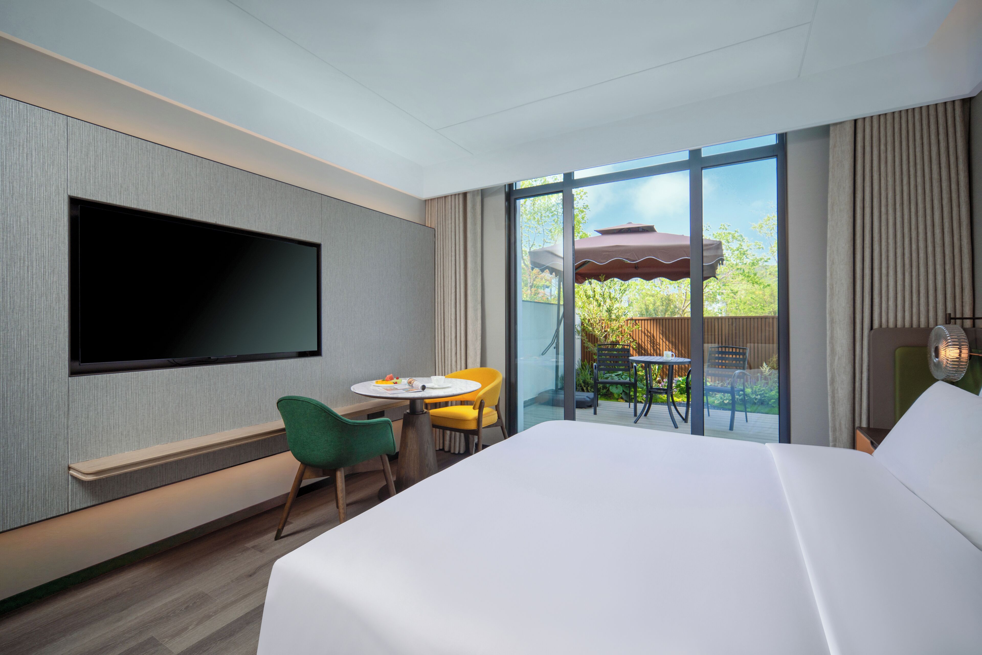 Habitación de hotel con una cama grande, televisor de pantalla plana en la pared, pequeña mesa de comedor con sillas, y puerta corrediza de vidrio que da a un patio exterior con mesa y sillas.