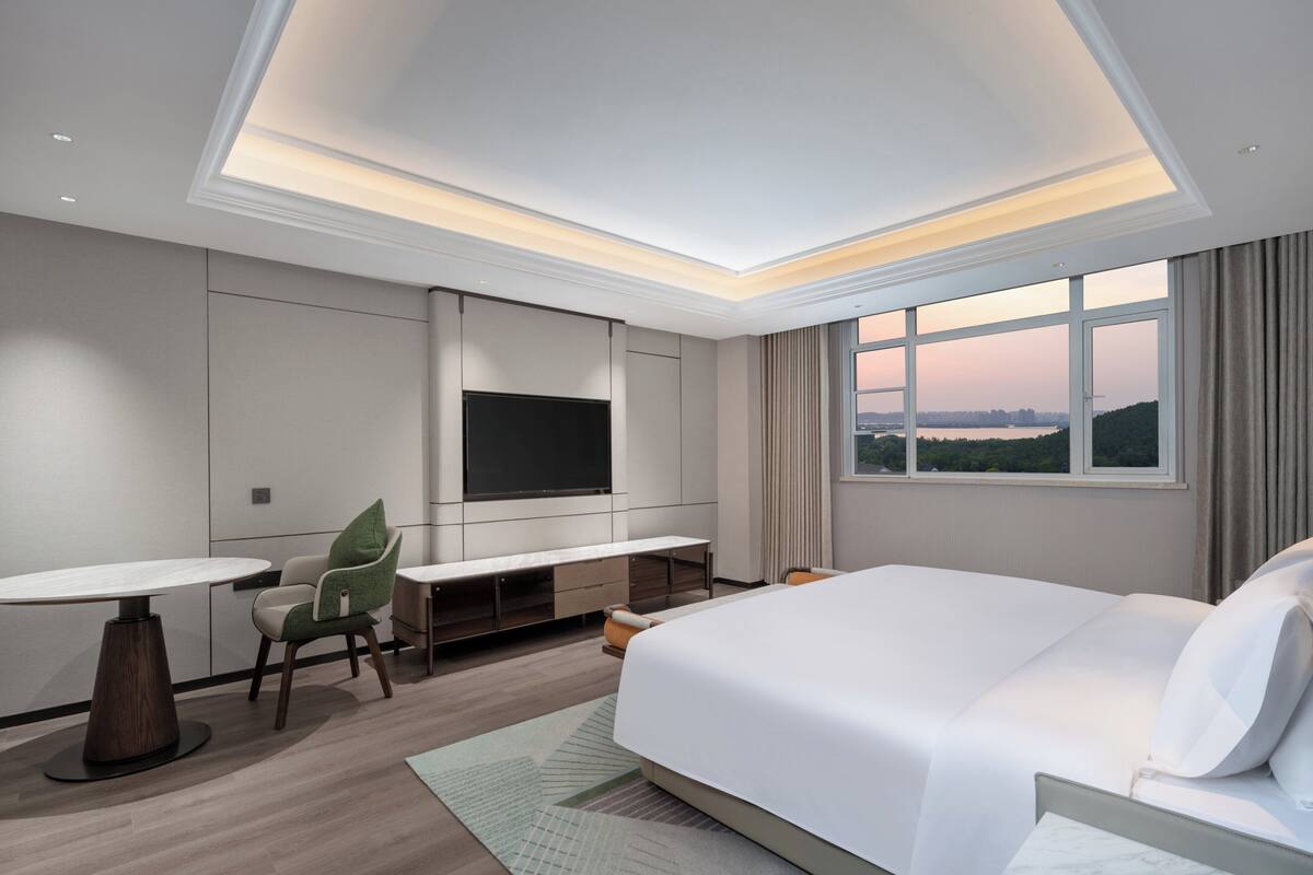 Ein modernes Hotelzimmer mit einem großen Bett, einem Schreibtisch, einem Sessel, einem an der Wand befestigten Fernseher und Blick auf die Stadt durch das Fenster.