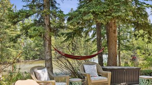 A hammock, wicker chairs, a fire pit, and a hot tub in a forested area with a view of a river. - The perfect place for family and friends to gather for a Rocky Mountain getaway! (Canmore)
