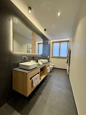 A modern bathroom with a large mirror, two sinks, a toilet, and a window.