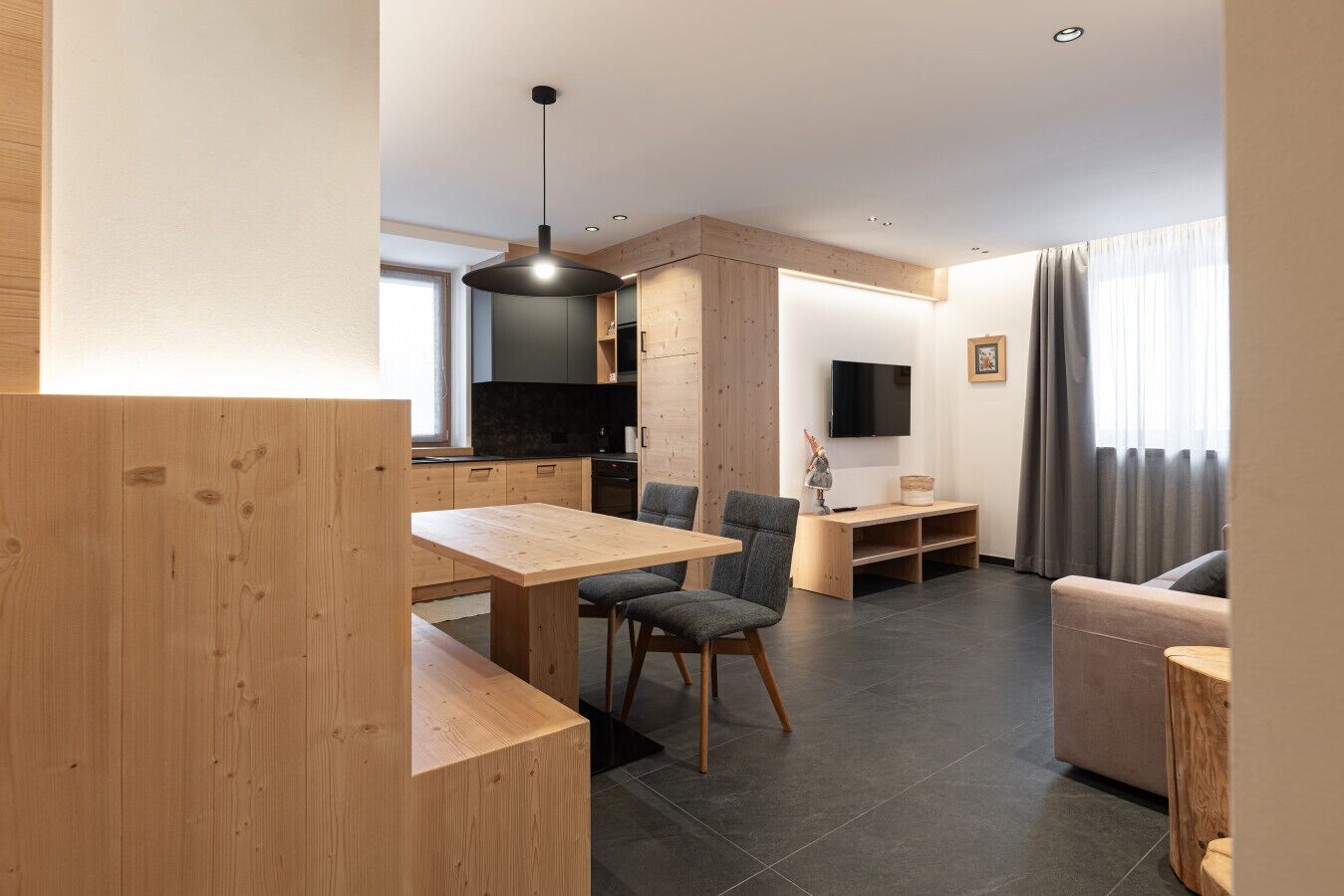 A modern living room with a wooden dining table, grey upholstered chairs, a grey sofa, and a TV mounted on the wall.