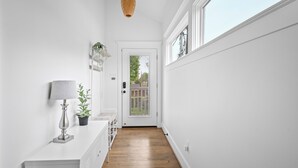 A narrow hallway with white walls, a wooden floor, a white console table with a lamp, and a door leading outside. - Solace One | Lindale Park Houston (Houston)