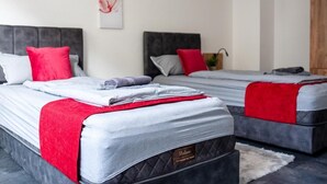 A hotel room with two beds, each with a grey mattress and red bedding. There is a red abstract artwork on the wall and a wooden wardrobe in the corner. - Livemore24 - Apartment 4 in Duisburg 24/7 check-in (Duisburg)