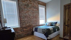 A bedroom with a brick wall, wooden flooring, a bed with a quilt, two windows with blinds, and a ceiling fan. - Charming 2-bedroom apartment in lovely Walnut Cove with AC, WiFi (Walnut Cove)