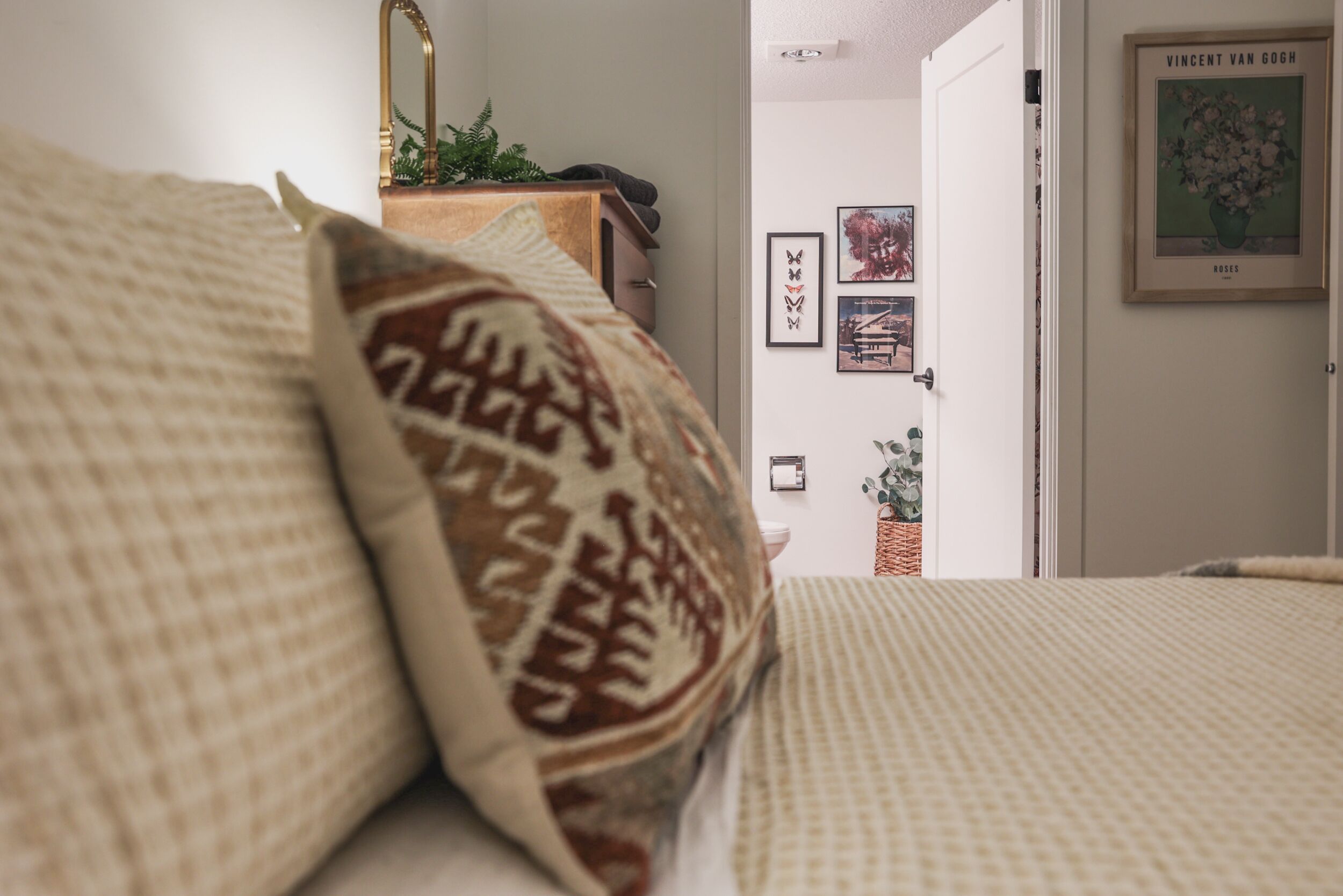 A bedroom with a bed, pillows, a dresser, a mirror, and a door leading to another room.