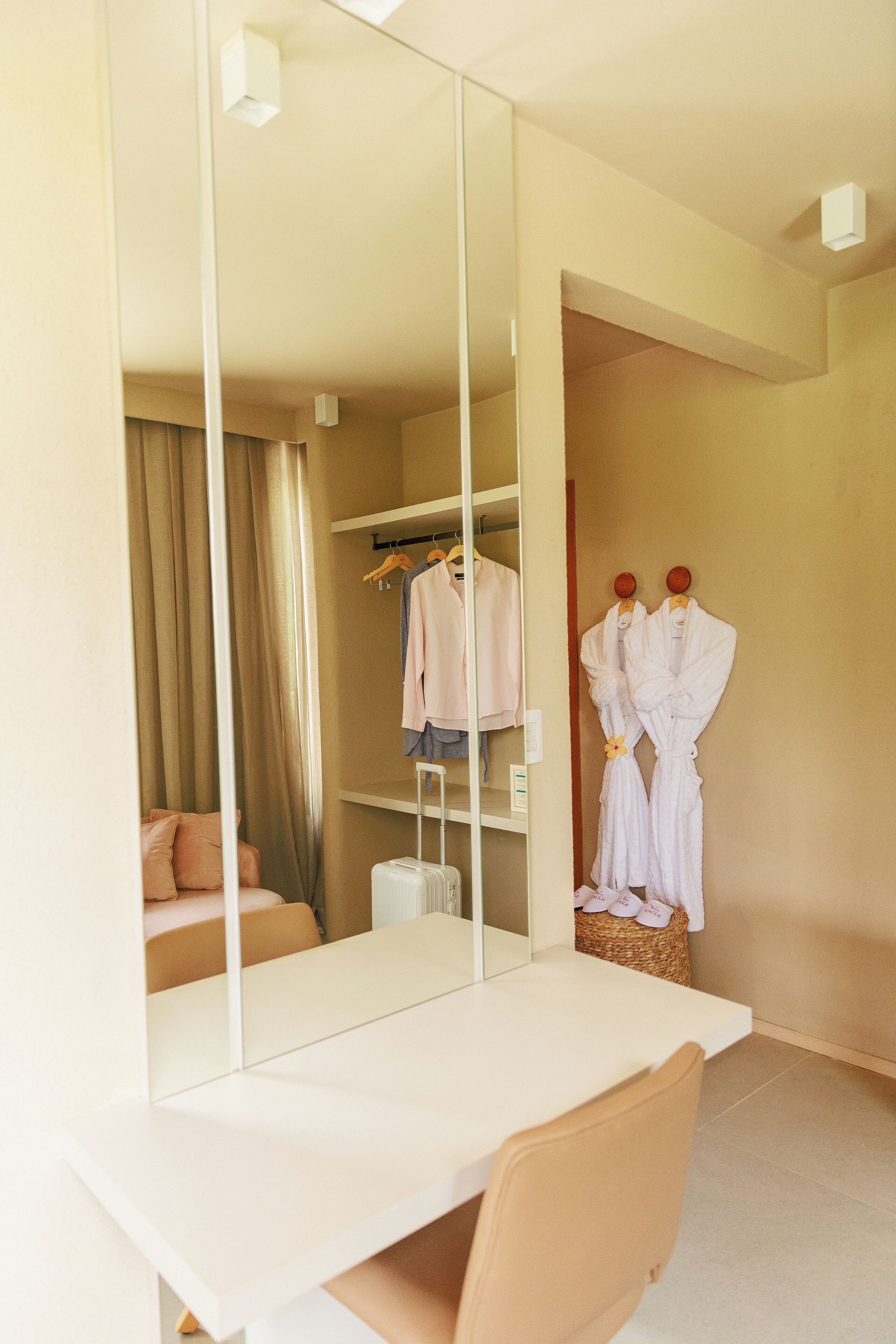 A hotel room with a large mirror, a desk, a chair, a wardrobe, and a hanging robe.