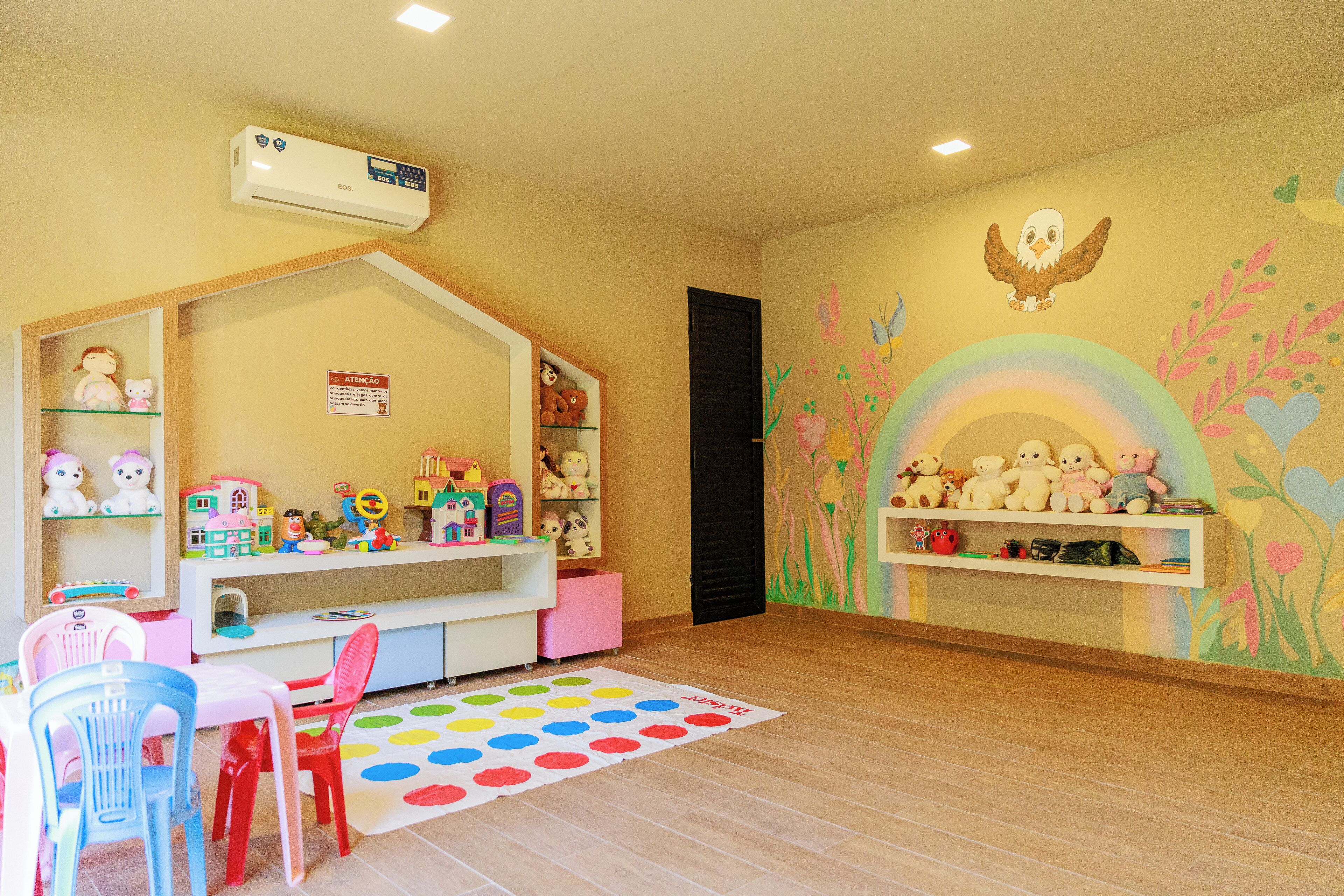 A children's playroom with a white playhouse, colorful toys, and a rainbow mural on the wall.
