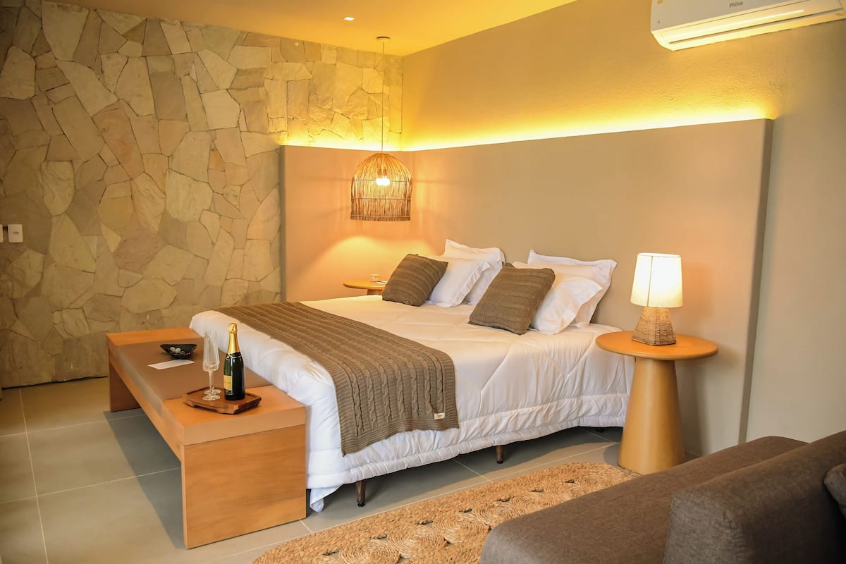 Ein Schlafzimmer mit Steinwand, einem Bett mit weißer Bettwäsche, einem Nachttisch mit Flasche und Glas sowie einer Lampe auf einem Beistelltisch.