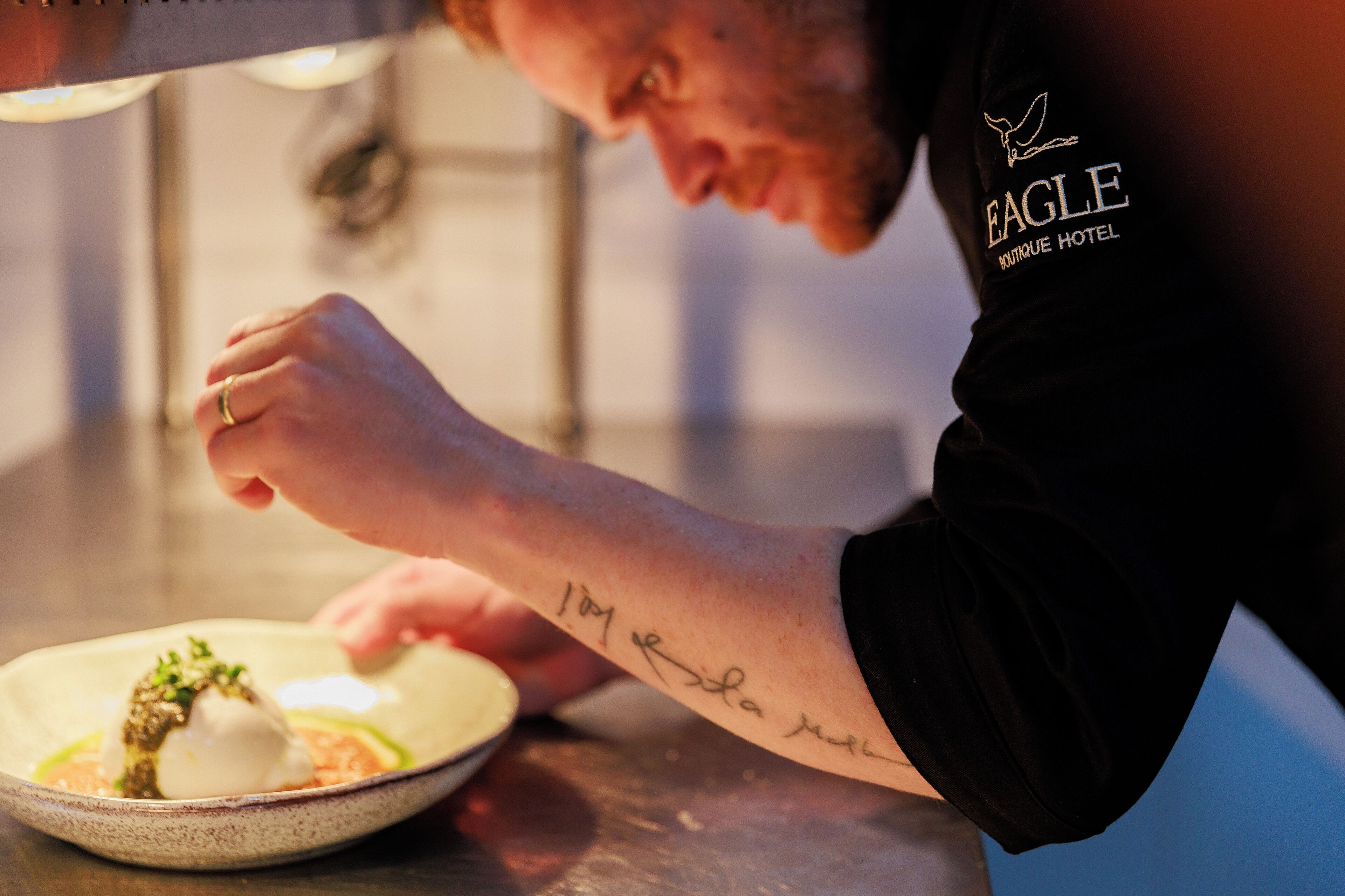 A person with a tattoo on their arm, wearing a black shirt with a logo, sitting at a table with a dish in front of them.