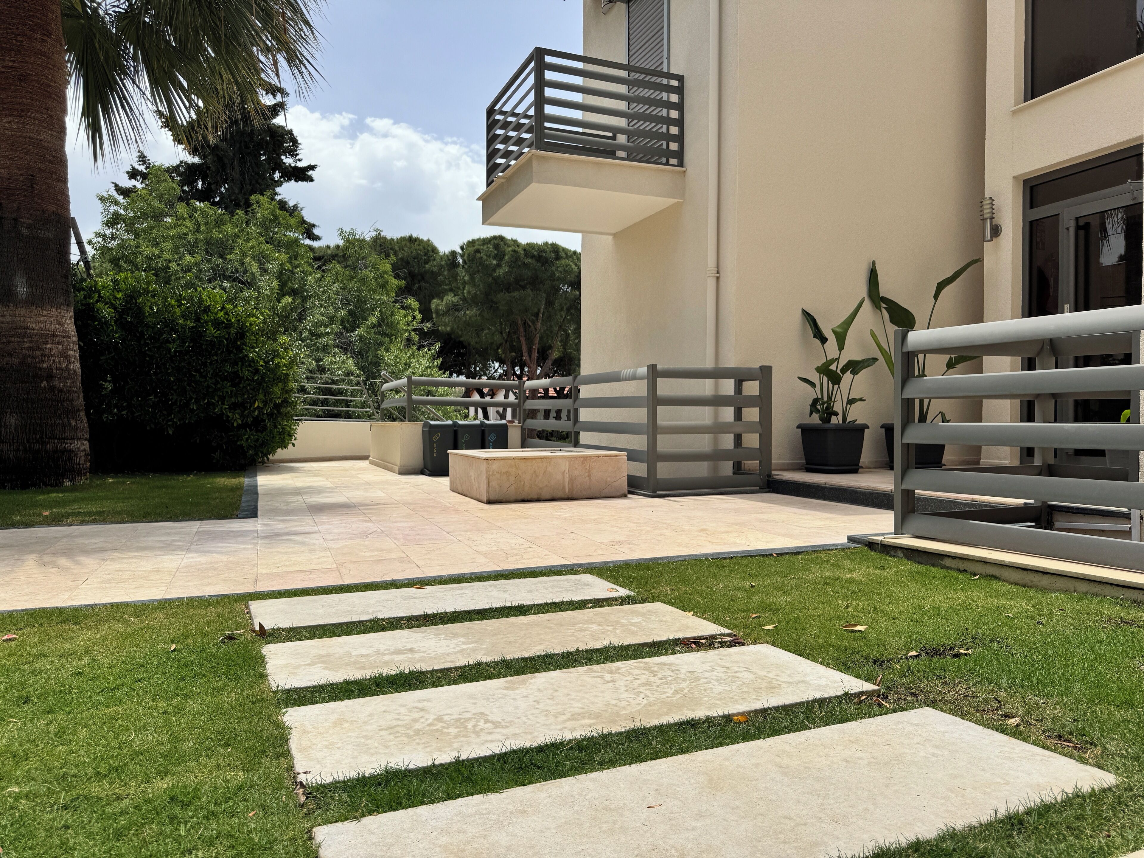 A modern outdoor patio with a stone pathway leading to a gated entrance, surrounded by greenery and a building with a balcony.