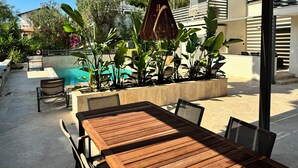Un área junto a la piscina con una mesa y sillas de madera, rodeada de plantas exuberantes y una estructura de pérgola.