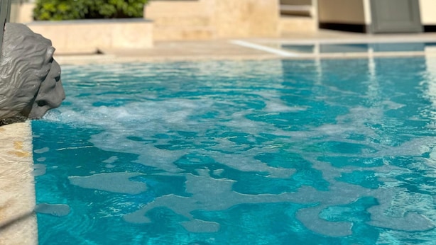 Una piscina con agua azul cristalina, rodeada por un edificio moderno con balcones y un jardín con árboles y arbustos.