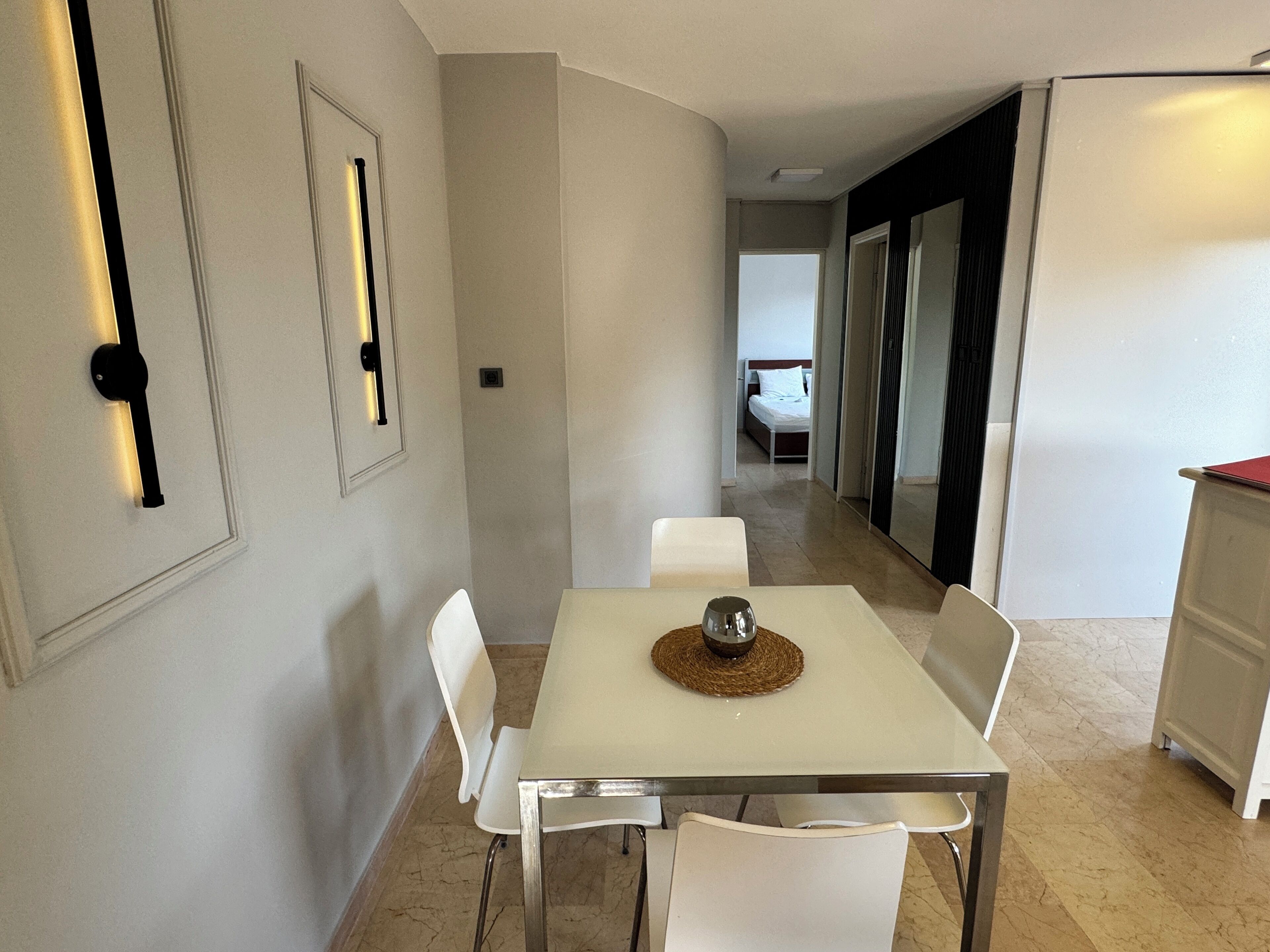 A hotel room with a dining area featuring a white table and chairs, a minibar, and a bed in the background.