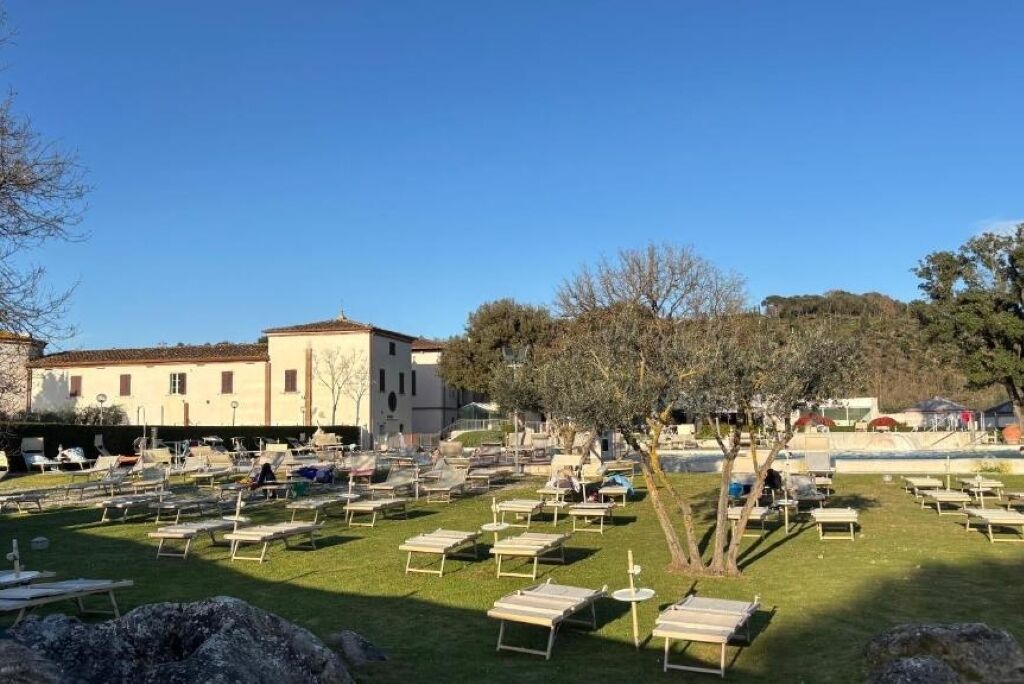 A courtyard with numerous sun loungers arranged in rows, a building with a tiled roof in the background, and trees providing shade.