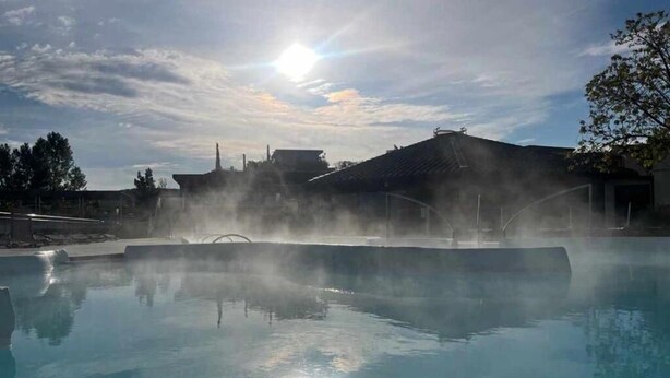Une piscine dotée d’un système de diffusion de vapeur, entourée de bâtiments et d’arbres, sous un ciel partiellement nuageux.