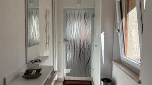 Une salle de bain moderne dotée d’une porte de douche en verre, d’un lavabo avec un miroir et d’une fenêtre laissant entrer la lumière naturelle.