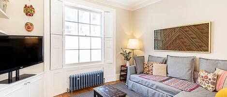 A living room with a large TV, a sofa, a coffee table, and a window with blinds.