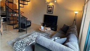 A modern living room with a curved gray sofa, a black spiral staircase, a flat-screen TV, and a patterned rug. - Condo Great for Large Family (Sandusky)
