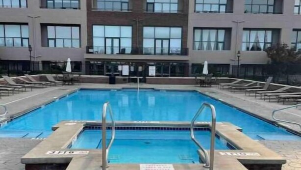 A rectangular swimming pool with a concrete deck and metal handrails, surrounded by lounge chairs and umbrellas, with a modern building in the background. - Condo Great for Large Family (Sandusky)
