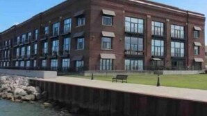 A multi-story brick building with large windows, a grassy area in front, and a waterfront. - Condo Great for Large Family (Sandusky)
