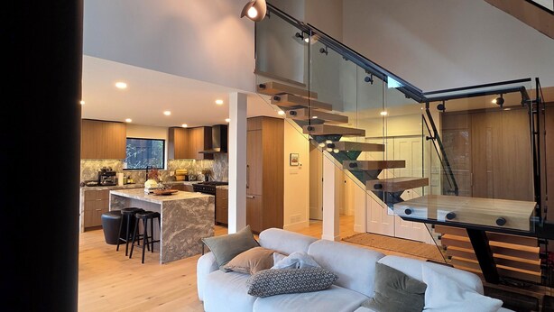 A spacious living area with a modern sofa, a kitchen in the background, and a staircase leading to an upper level.