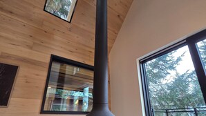 A modern interior with a large chimney, wooden ceiling, and large windows offering views of trees.