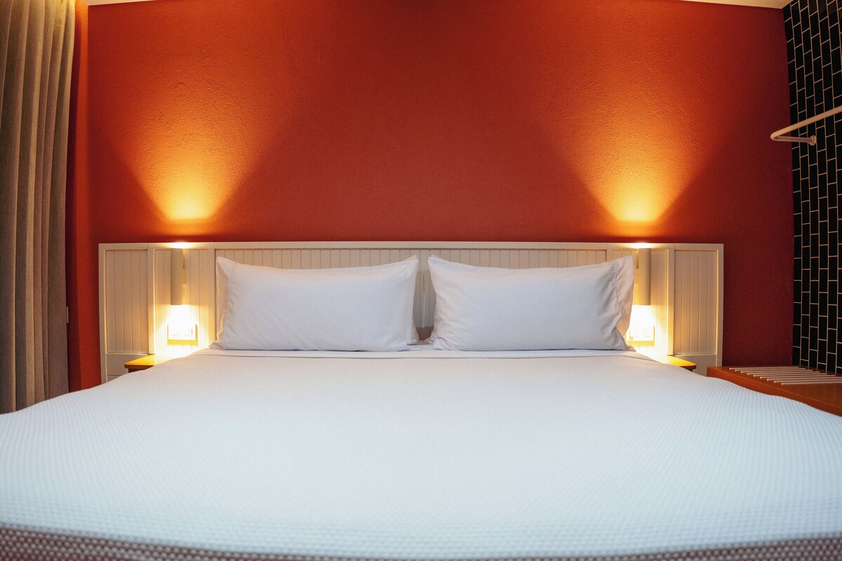 A large bed with white linens, two bedside lamps, and a red wall.