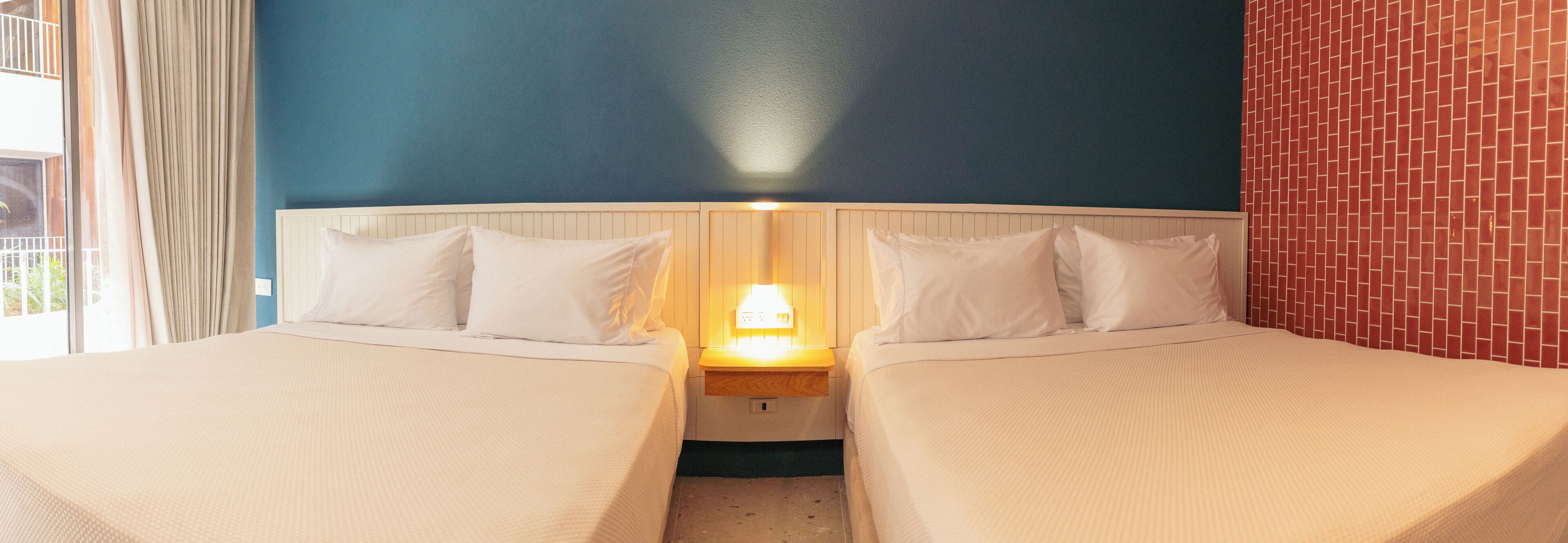 Quarto de hotel com duas camas, mesa de cabeceira com abajur, janela com cortinas e parede com padrão de tijolos.