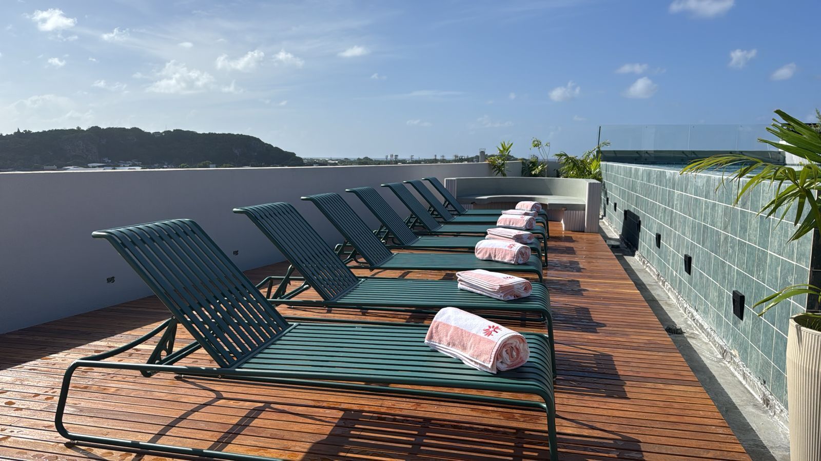 A row of green lounge chairs with towels on a wooden deck overlooking a body of water and a distant hill.